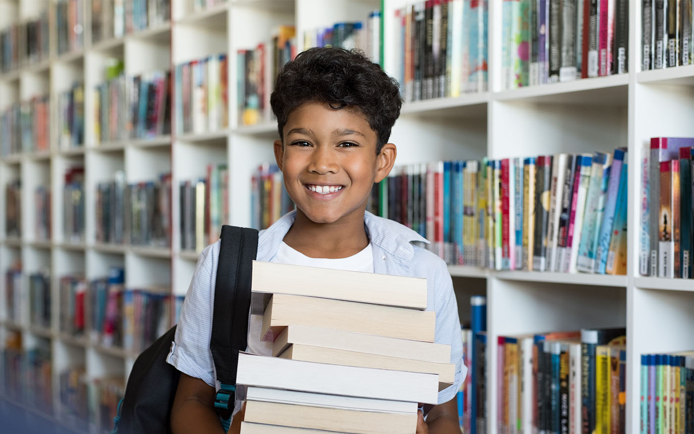 A smiling boy in the library holding a large stack of books of his chest.