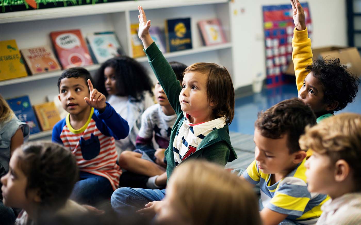 A group of multi-ethnic Kindergarten children gathered on the carpet a few children raising hands among them one with an excited expression to answer a teacher prompt.