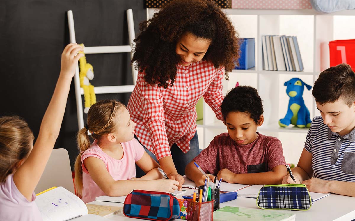 Homeschool teacher assisting a small group of learners with their work at a table inside a cheery room with shelf unit.