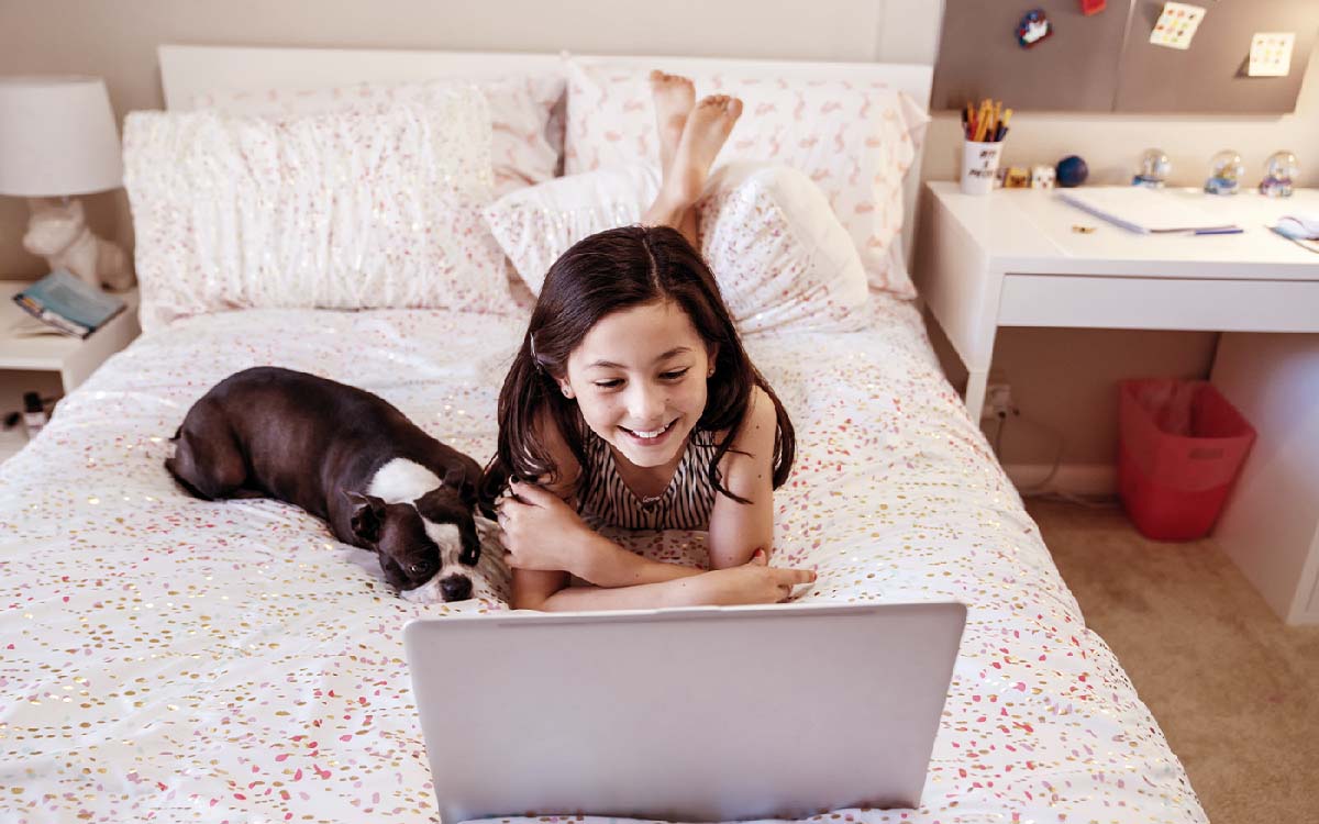 Smiling happy girl laying on bed with Boston Terrier home schooling on laptop computer.