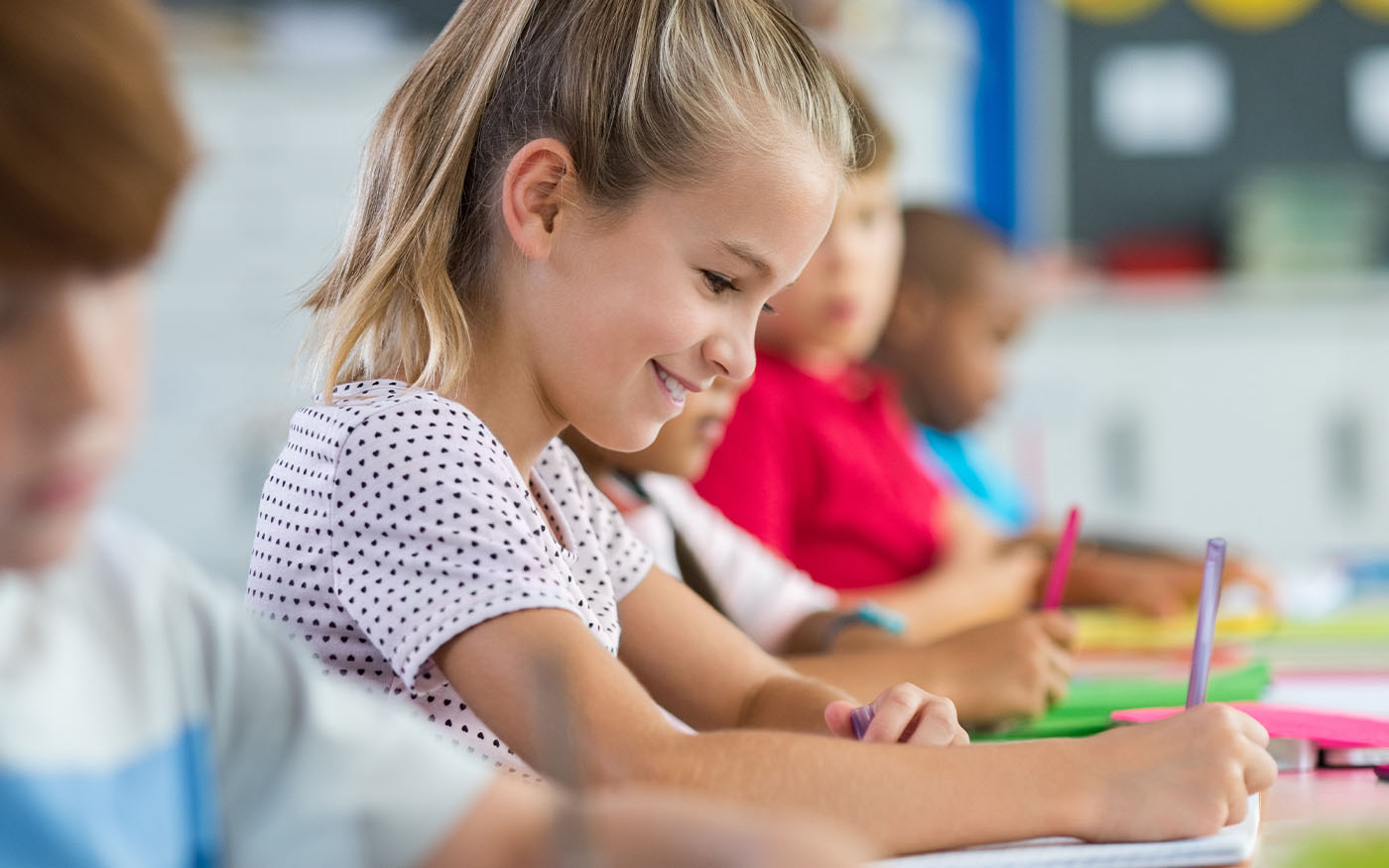 Elementary student at desk looking down while writing and smiling.