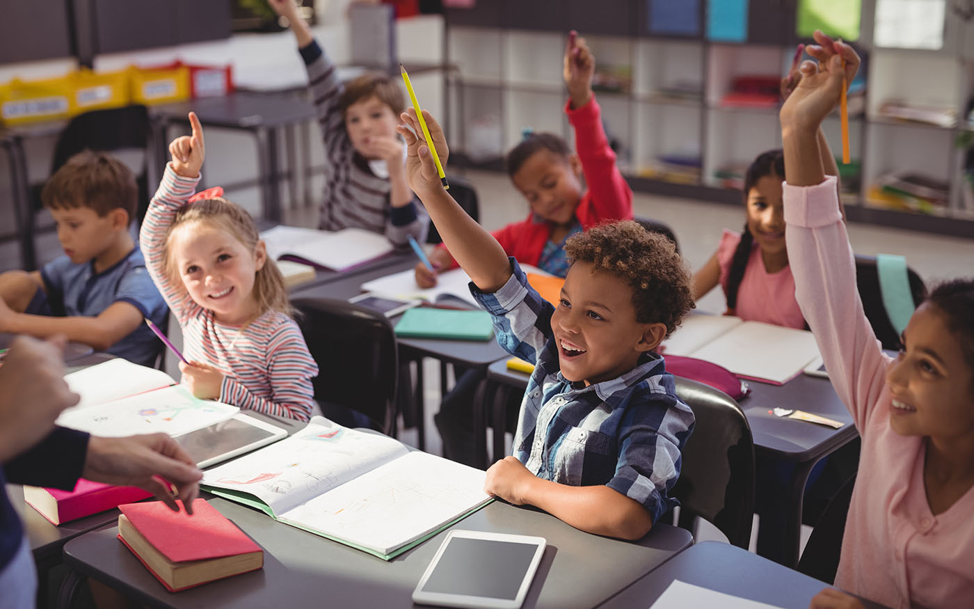 Students in a classroom sitting at their desks raising their hands excitedly.