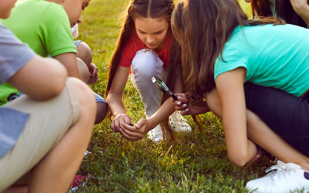 Curious homeschool group of middle school children explore nature and use loupe to study insects in grass.