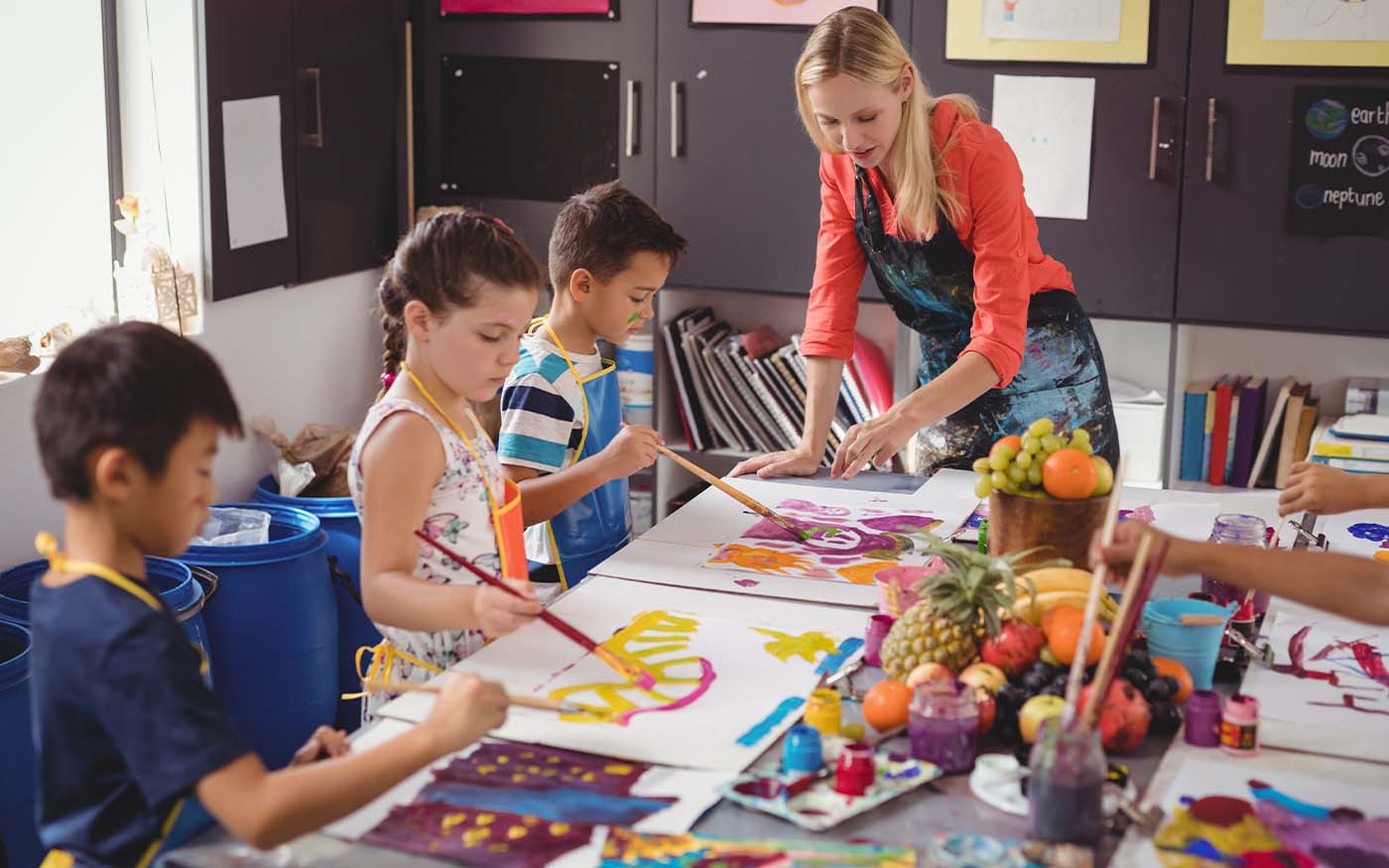 Small group of multi-ethnic primary students in an art lesson standing around a table while painting pictures with instructor looking on.