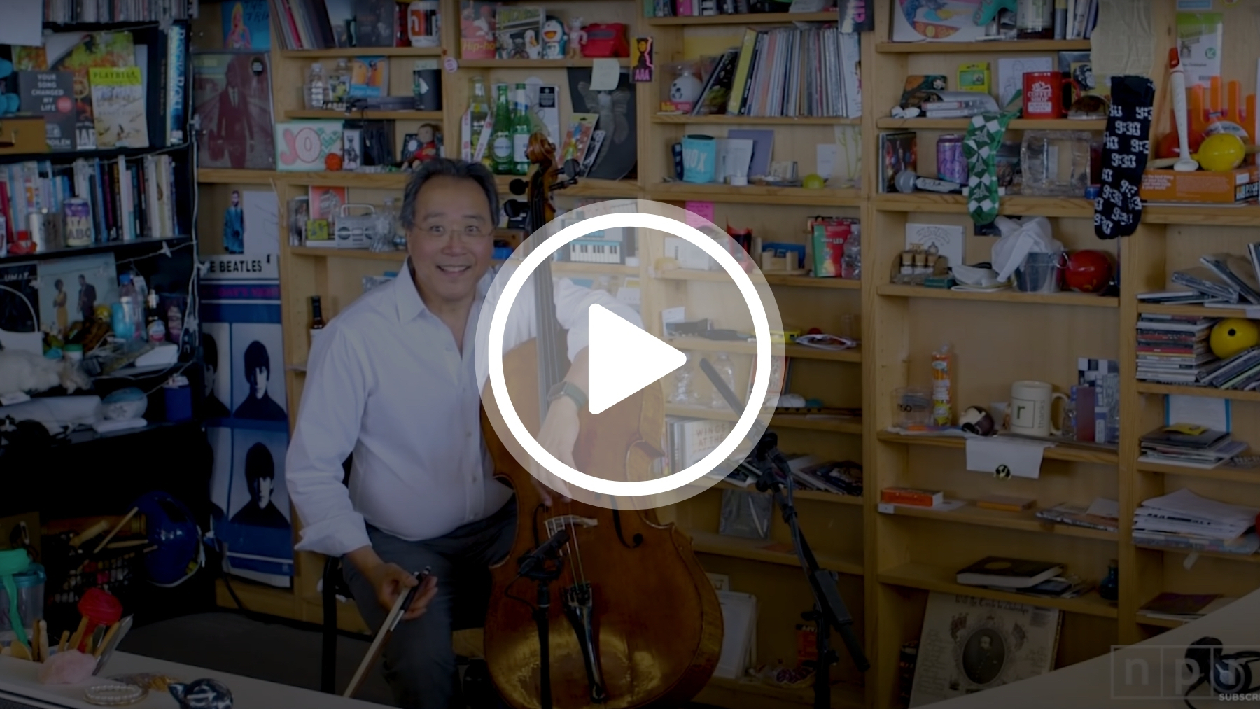 Yo-Yo Ma sitting in a chair surrounded by floor to ceiling shelves in the office of NPR's tiny desk concert holding his cello.