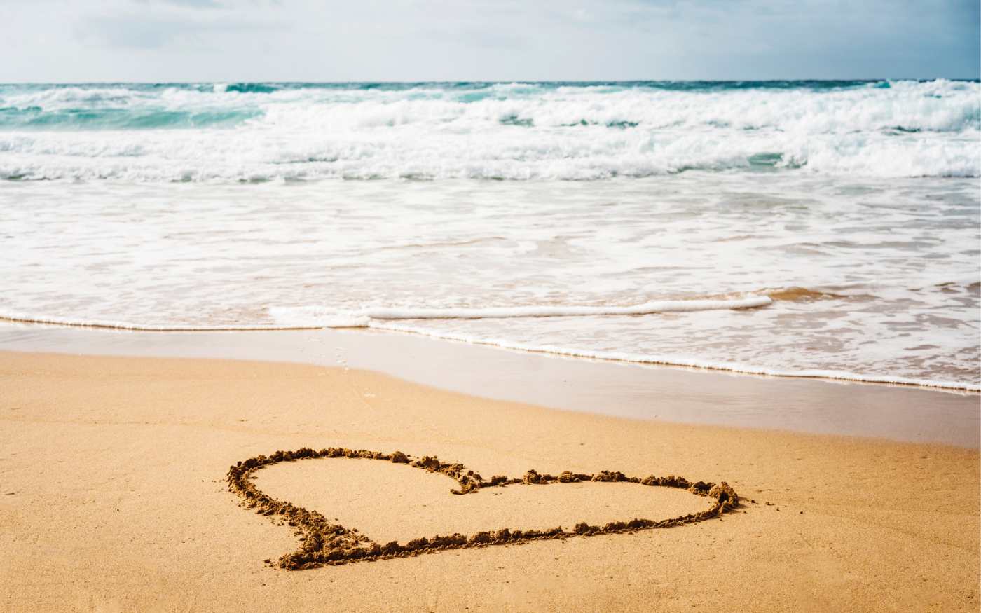 Image of beach sand by the ocean, with a heart shape drawn in the foreground in the sand.